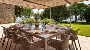 Un espace de restauration en plein air avec une table dressée pour six personnes, entourée de chaises en osier, sous une pergola offrant une vue sur un jardin luxuriant et un plan d’eau.