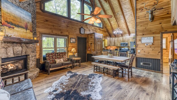 Een rustiek houten blokhutinterieur met een stenen open haard, een houten eettafel en een groot raam met uitzicht op het bos.