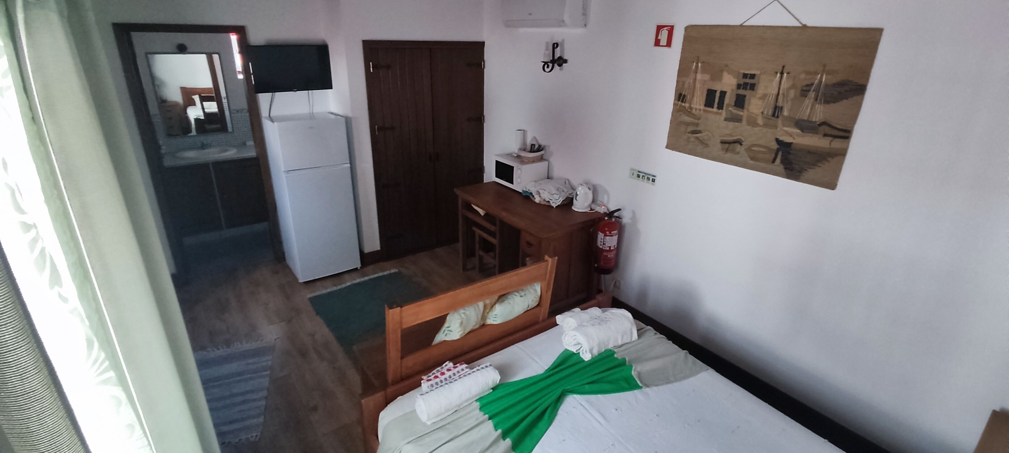 Quarto de hotel com cama, mesa de cabeceira de madeira, pequena mesa, espelho, frigorífico branco, porta de madeira, extintor de incêndio e quadro pendurado na parede.