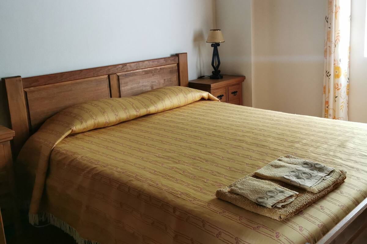 Een net opgemaakt bed met een beddekleed met dessin, twee opgevouwen handdoeken, een bedlampje en een wandlamp.