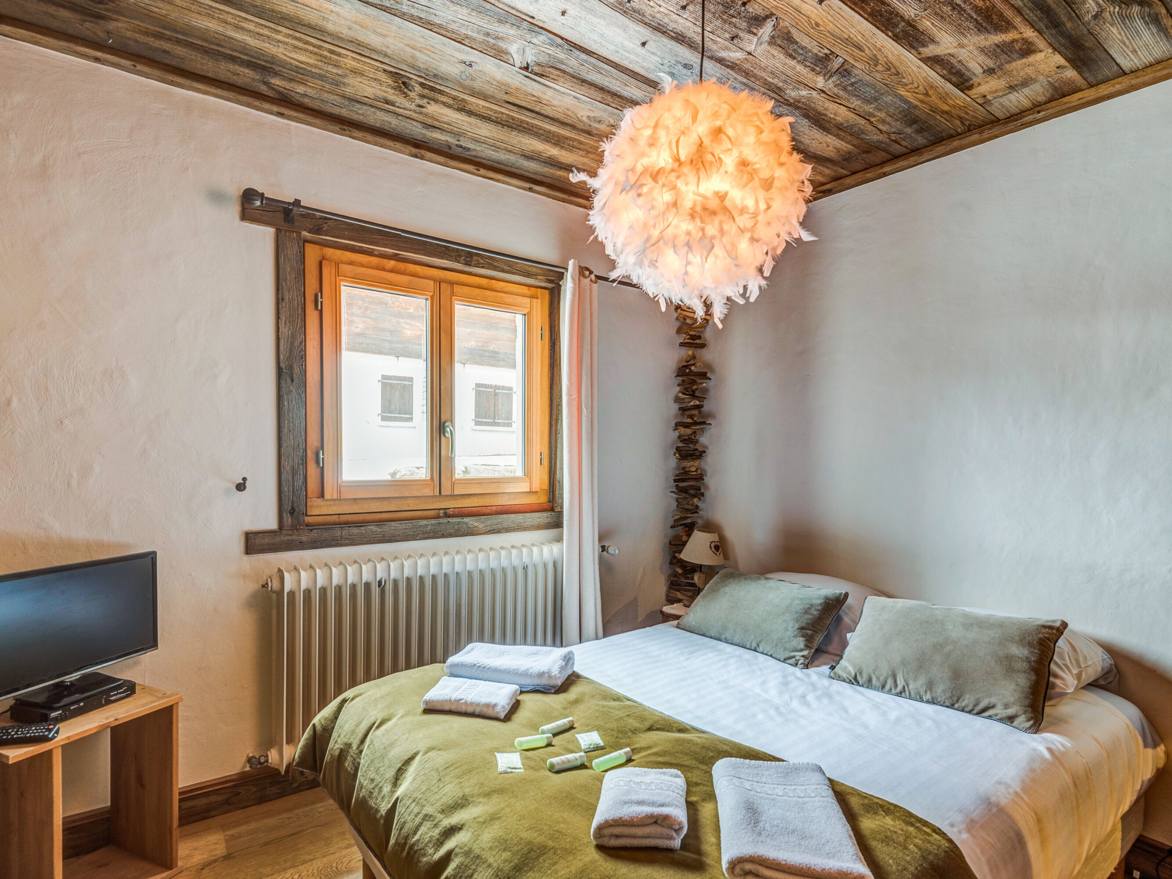 Een slaapkamer met een bed, een televisie op een houten standaard, een raam met houten luiken en een radiator.