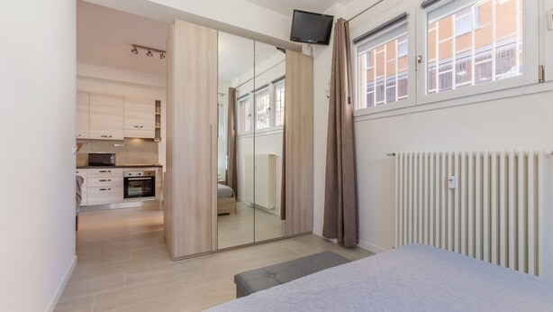 Ein modernes Zimmer mit einem Bett, einem Spiegel, einem Fernseher und einer Küche im Hintergrund.