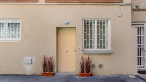 Ein Gebäude mit einer beigefarbenen Fassade, einer gelben Tür, zwei Fenstern mit weißen Rahmen und zwei Blumentöpfen.
