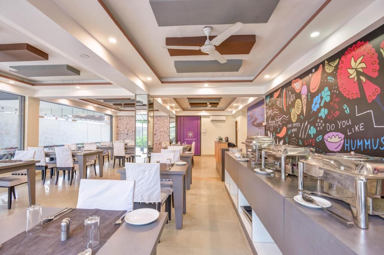 A modern dining area with tables and chairs, a buffet counter with food containers, and a mural with colorful illustrations on the wall.