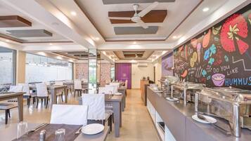 Ein moderner Essbereich mit Tischen und Stühlen, eine Buffettheke mit Speisebehältern und ein Wandgemälde mit farbenfrohen Illustrationen.