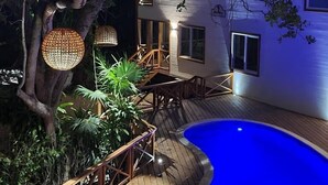 Une terrasse en bois avec une piscine, entourée d’une végétation luxuriante et d’un bâtiment blanc doté de fenêtres et d’une porte.