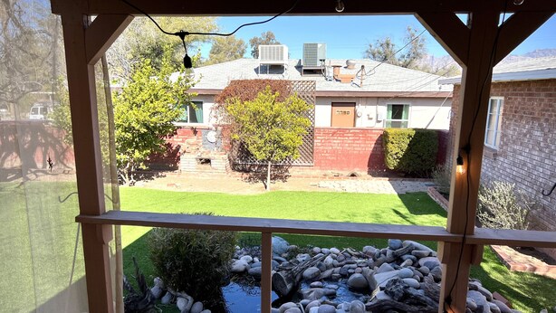 Ein überdachter Außenbereich mit Blick auf einen Garten, einen kleinen Teich und ein Backsteinhaus.