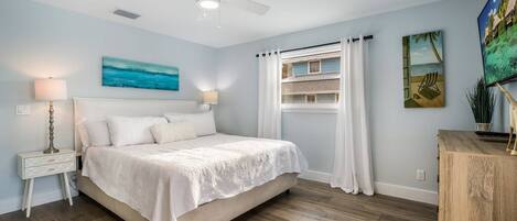 A bedroom with a bed, nightstands, a ceiling fan, a window with curtains, and a painting on the wall.
