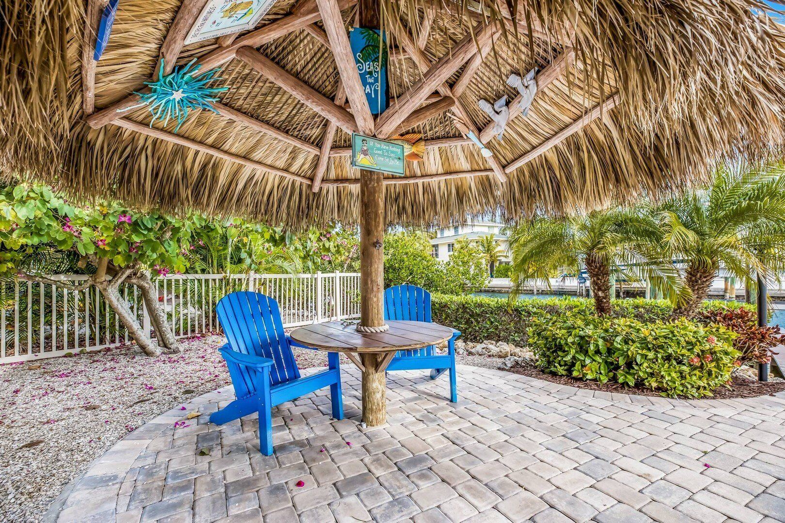 A thatched-roof outdoor seating area with two blue chairs and a table, surrounded by greenery and a white fence.