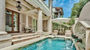 A pool area with steps leading into the water, a covered seating area with umbrellas, and a stone wall with a water feature. - Lady Martinique | Exquisite 5 Bedroom with Private Pool, Outdoor Fireplace (Rosemary Beach)