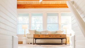 A staircase leading to a loft bedroom with a wooden ceiling, a bed, and a view of the outside through large windows. - Cozy cottage nestled in forest on BHI. Close to east beach and located in tranquil Sumners Crescent (Bald Head Island)