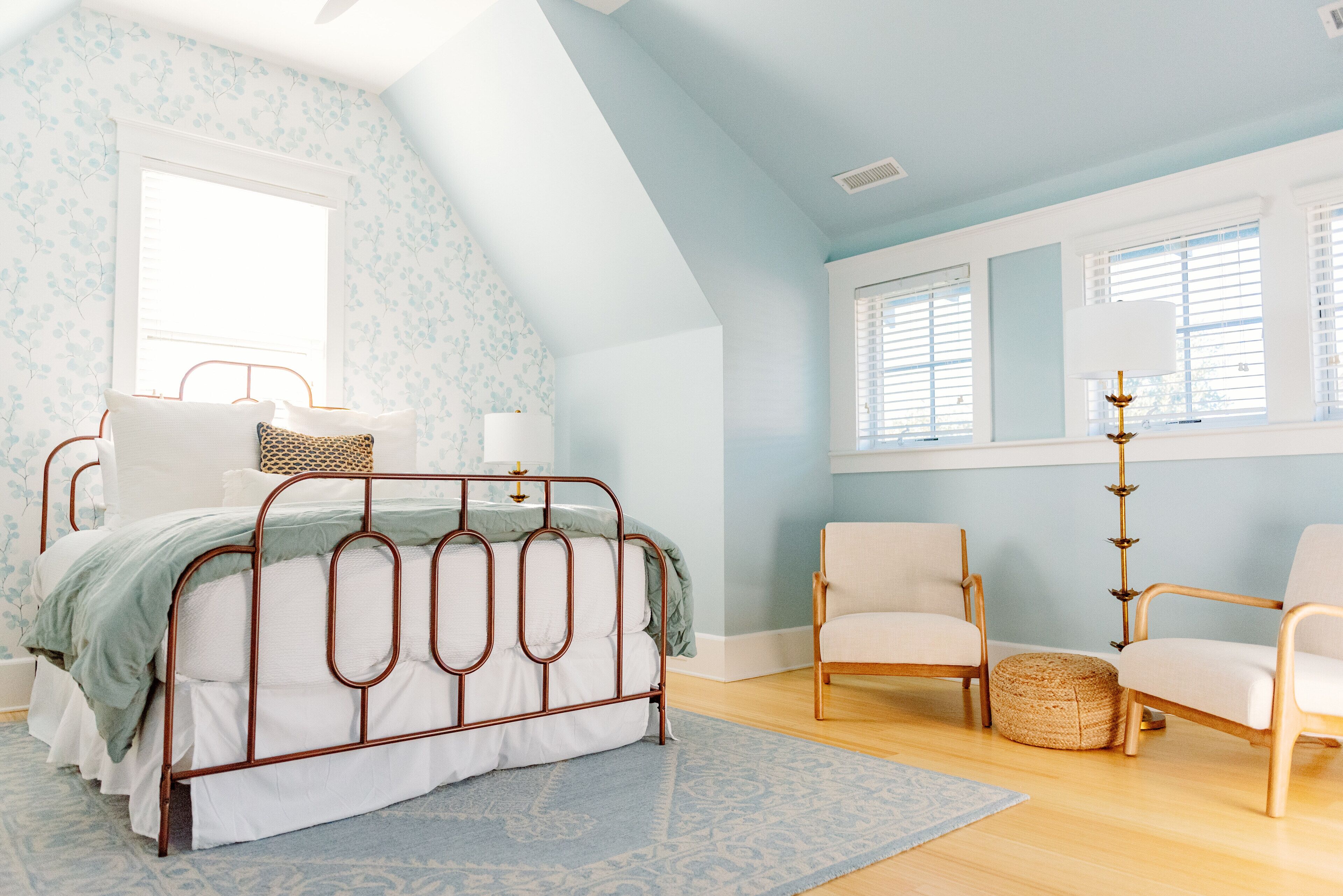 A bedroom with a sloped ceiling, a metal bed frame, two chairs, a bedside table, and a large window with blinds.