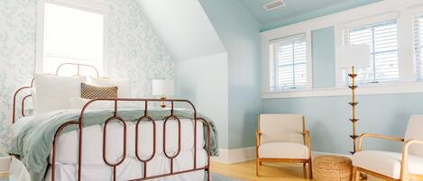 A bedroom with a sloped ceiling, a metal bed frame, two chairs, a bedside table, and a large window with blinds.