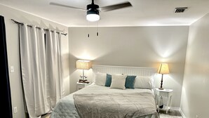 Un dormitorio con una cama, dos mesitas de noche con lámparas, un ventilador de techo y una ventana con cortinas.