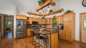Une cuisine avec des meubles en bois, un réfrigérateur en acier inoxydable, un îlot central avec un comptoir en granit et des tabourets de bar, ainsi que des luminaires suspendus.