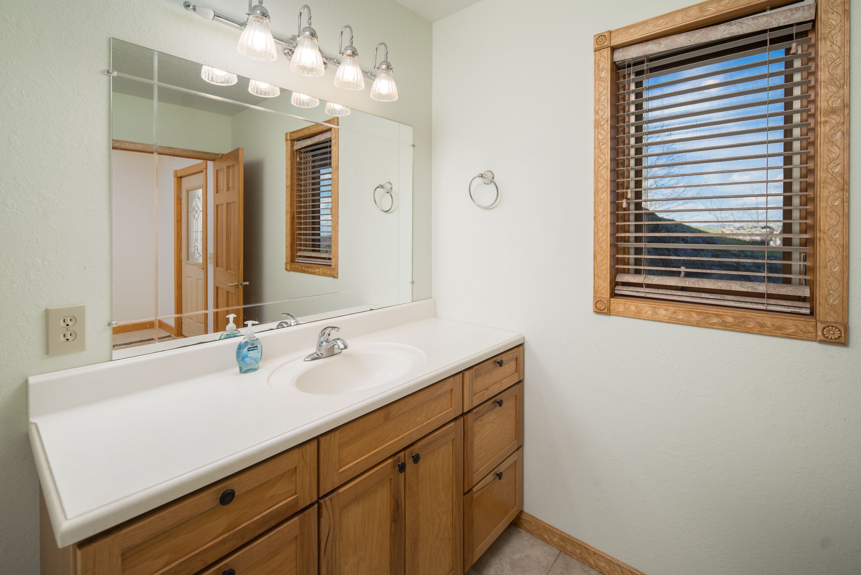 Un baño con un espejo grande, armarios de madera, un lavabo blanco y una ventana con persianas.