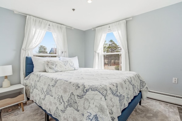 Een slaapkamer met een bed, gordijnen, een nachtkastje en een raam met uitzicht.