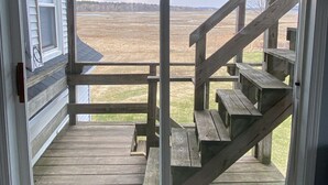 Eine Holzterrasse mit einer Treppe, die nach draußen führt, Blick auf ein grünes Feld und ein Gebäude im Hintergrund.