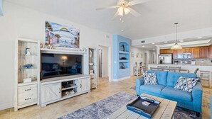 Ein geräumiges Wohnzimmer mit Fernseher, Sofa und Couchtisch, Deckenventilator sowie einer Küche im Hintergrund.