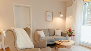 A modern living room with a beige sofa, a small round table, and a chair. There is a door and a window with curtains. - The vacation apartment with three terraces is located between the forest and the lake (Ratzeburg)