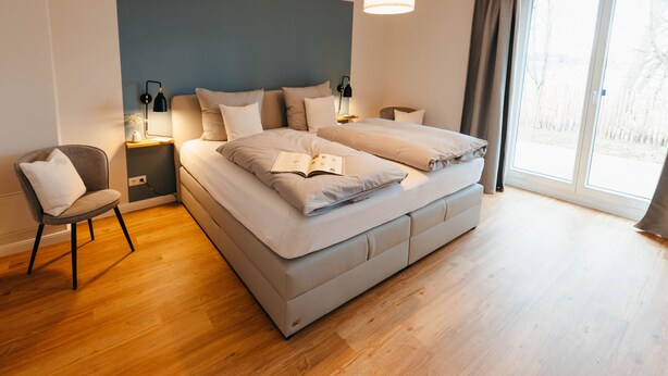 A modern bedroom with a large bed, bedside tables, a reading chair, and a large window. - The vacation apartment with three terraces is located between the forest and the lake (Ratzeburg)
