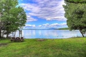 A private beach with a wooden bench and a sign, surrounded by lush green trees and a calm lake under a partly cloudy sky. - Beach House Chalet - shared access to Rangeley Lake (Rangeley)