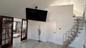 A spacious living area with a flat-screen TV mounted on the wall, a wooden floor, and a staircase leading to an upper level.