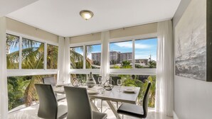 Ein stilvolles Esszimmer mit einem großen Tisch, vier Stühlen und Blick auf Palmen und Gebäude durch große Fenster.