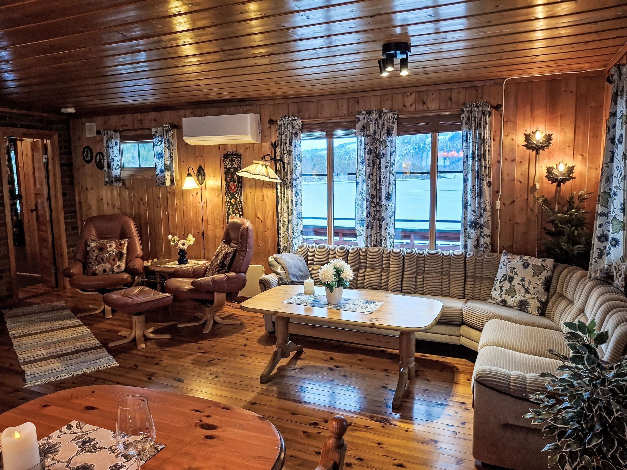 A cozy cabin interior with wooden walls and floors, a leather armchair, a sofa, a dining table, and a view through large windows.