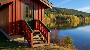 A cabin with a green door and red exterior, situated by a calm lake with a forested hill in the background. - Midtholmen by Interhome (Konsmo)