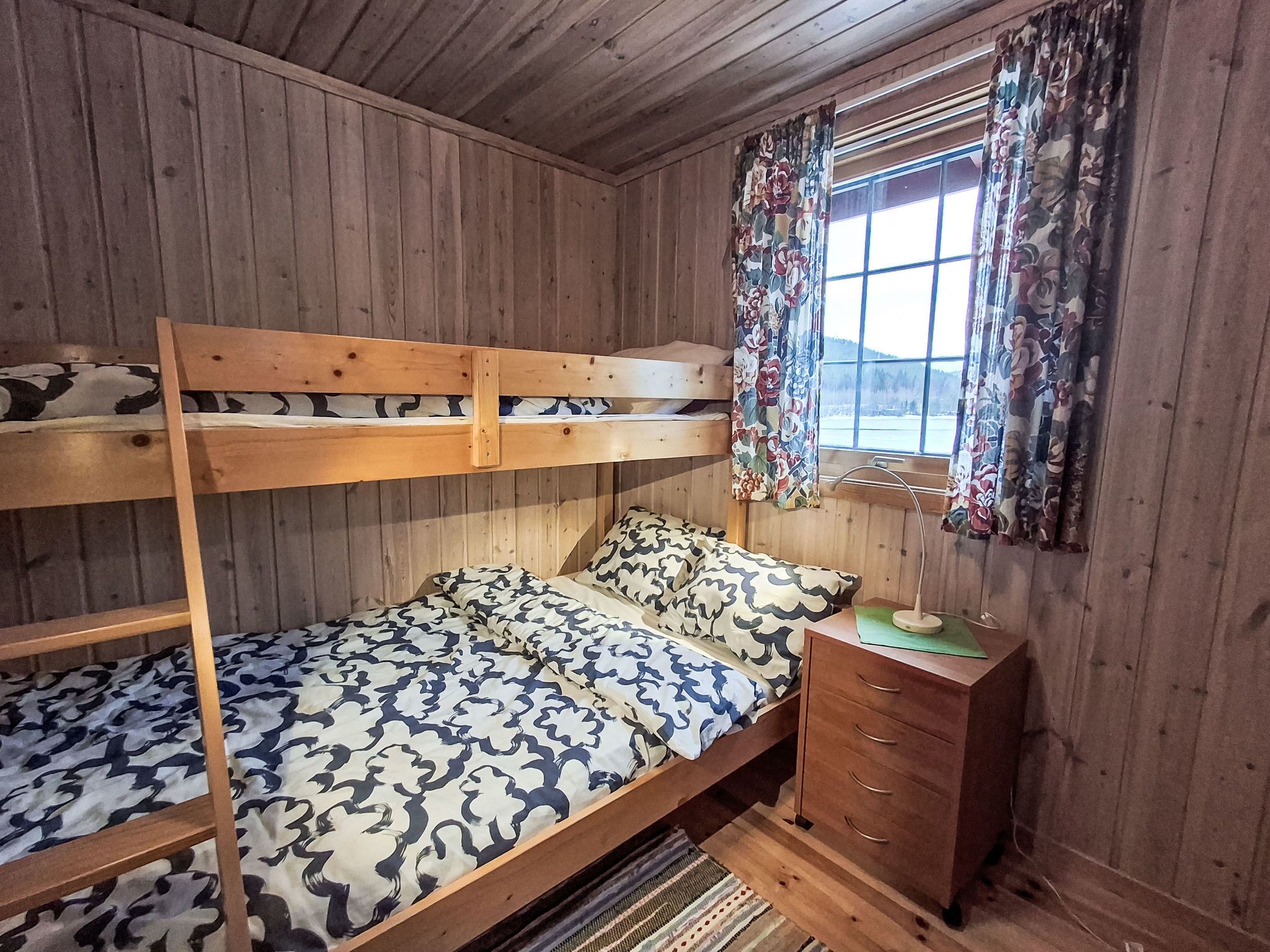 Ein Etagenbett mit gemusterten Betttextilien, eine Holzkommode mit Schubladen, ein Fenster mit floralen Vorhängen und Blick auf eine verschneite Landschaft.