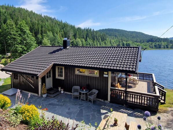 A wooden cabin with a tiled roof, a deck, and a view of a lake and forest.