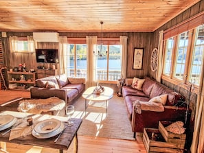 A cozy cabin interior with a wooden ceiling, a television, a sofa, a dining table set, and a view of a lake through large windows. - Staksholmen by Interhome (Konsmo)