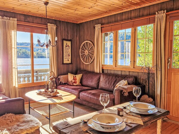 A cozy cabin interior with a leather sofa, wooden dining table, and large windows overlooking a lake. - Staksholmen by Interhome (Konsmo)