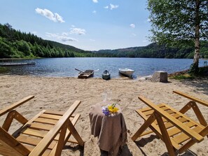 A beach with wooden chairs and a table set for a meal, overlooking a lake with boats and forested hills. - Staksholmen by Interhome (Konsmo)