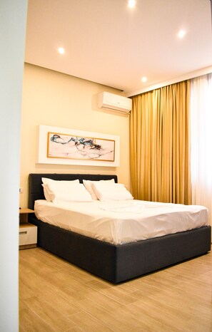 Quarto de hotel moderno com uma cama grande, mesa de cabeceira, ar condicionado fixado na parede e uma pintura abstrata emoldurada.