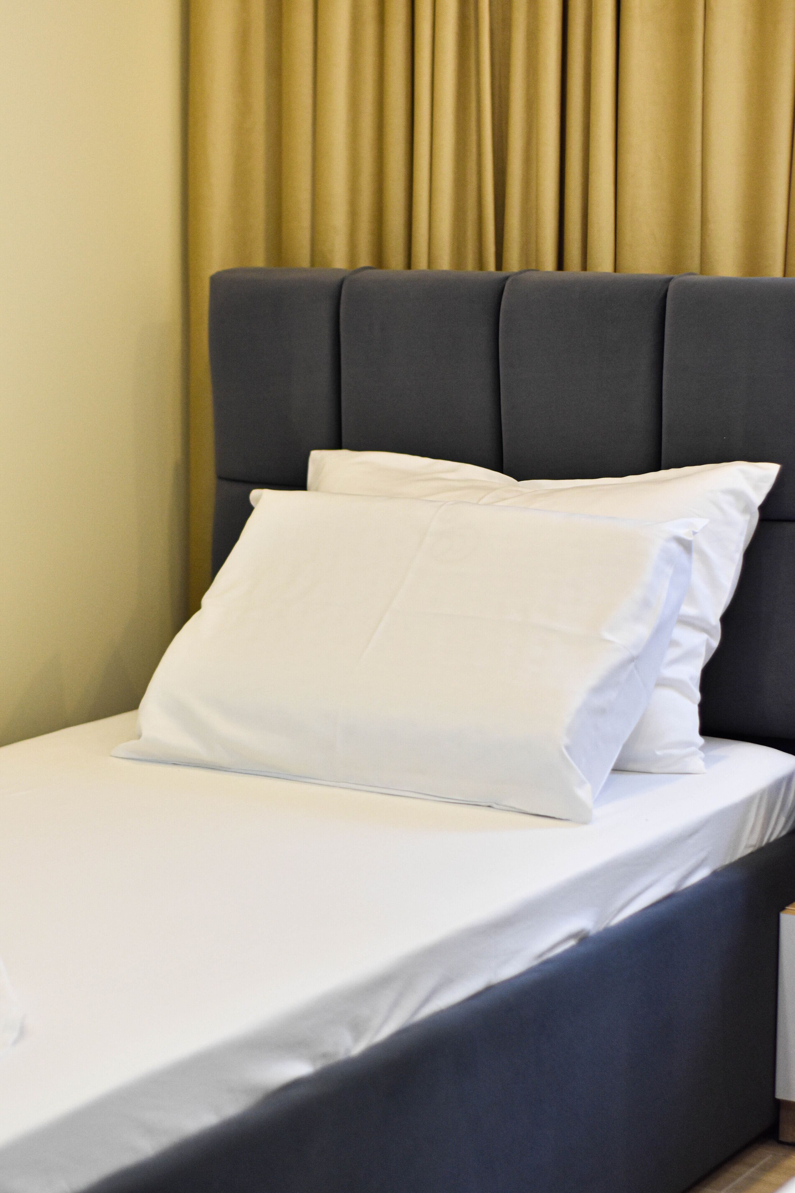 Una cama con cabecera oscura, ropa de cama blanca y una cortina beige al fondo.
