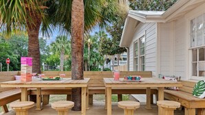 Eine Terrasse aus Holz mit mehreren Tischen und Bänken, umgeben von Palmen und einem Haus mit weißem Putz.