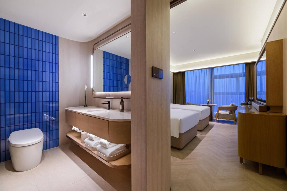A modern hotel room with a large bed, a bathroom with a vanity, and a view of the city through large windows.