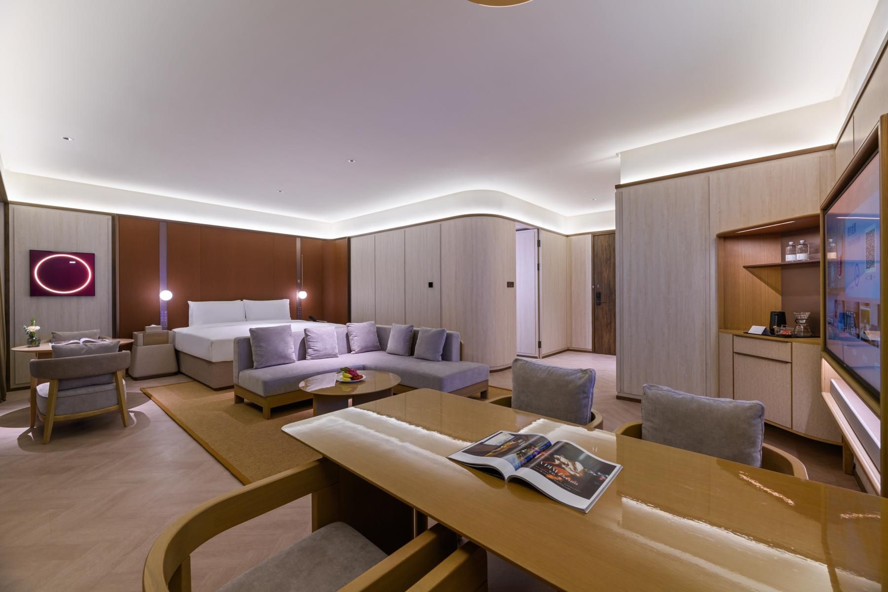 A modern hotel room with a bed, sofa, and armchairs, a dining table with magazines, and a TV mounted on the wall.