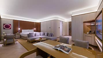 A modern hotel room with a bed, sofa, and armchairs, a dining table with magazines, and a TV mounted on the wall.