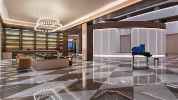 A spacious lobby with a checkered floor, a grand piano, a seating area with sofas, and a wall with illuminated shelves.
