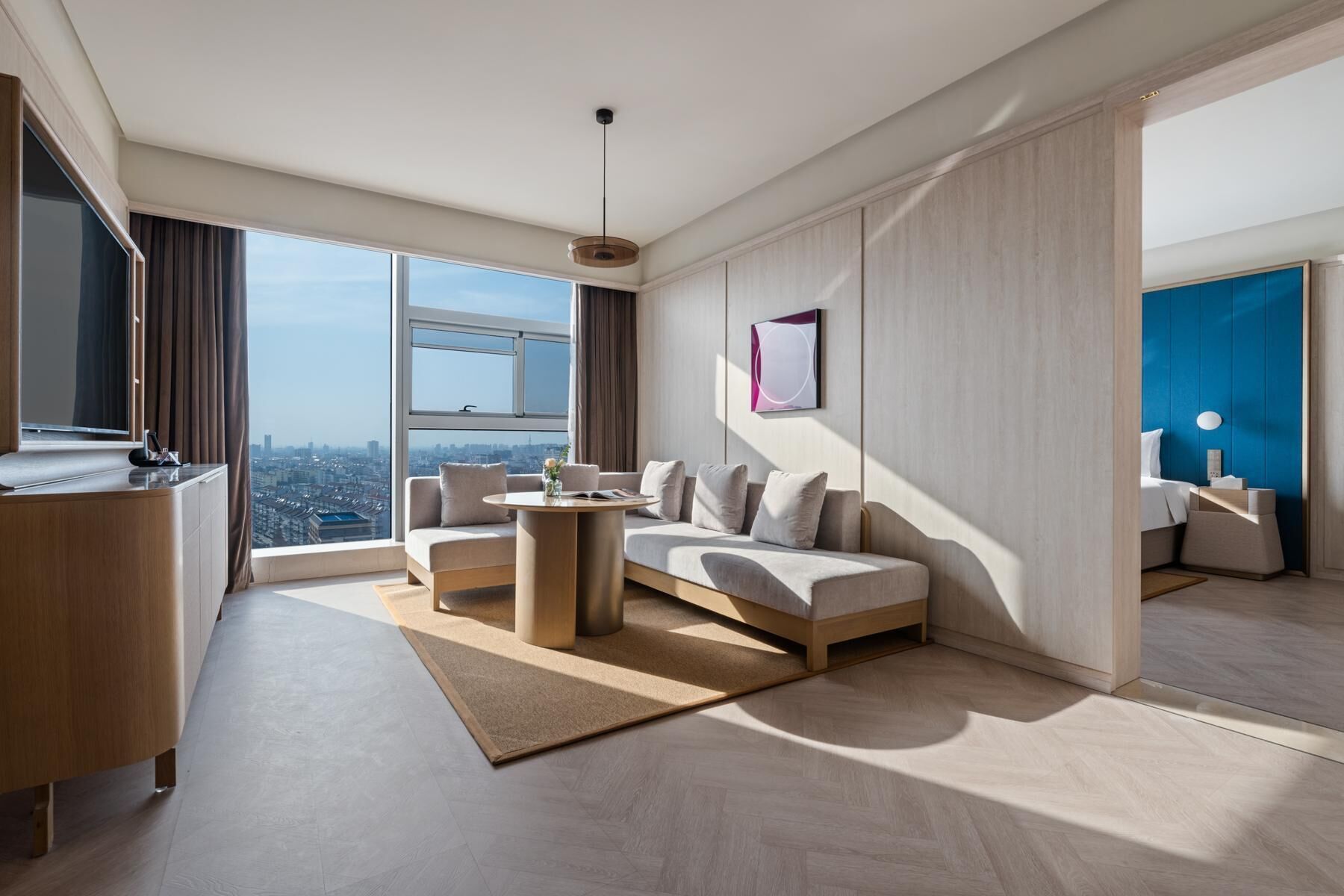 A modern hotel room with a large window, a sofa, a small table, and a desk.