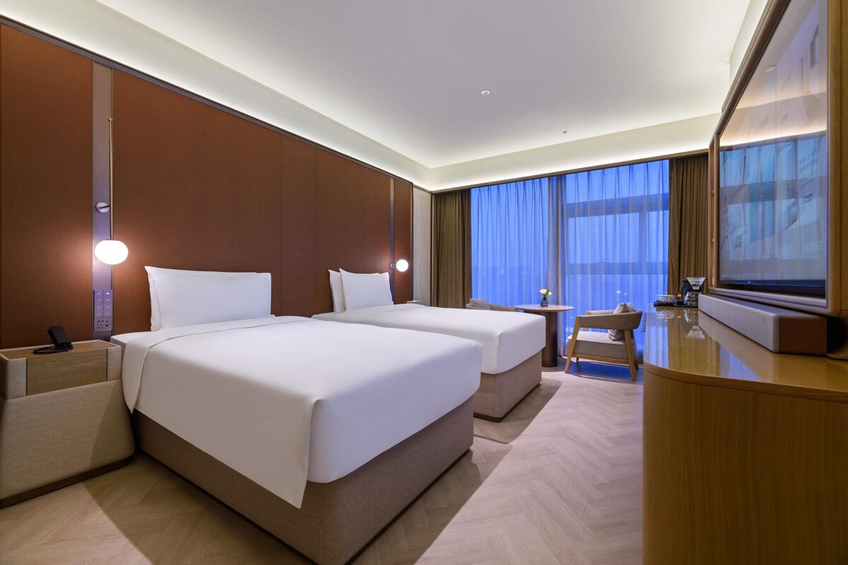 A hotel room with two beds, a desk, a chair, a television, and a large window with curtains.