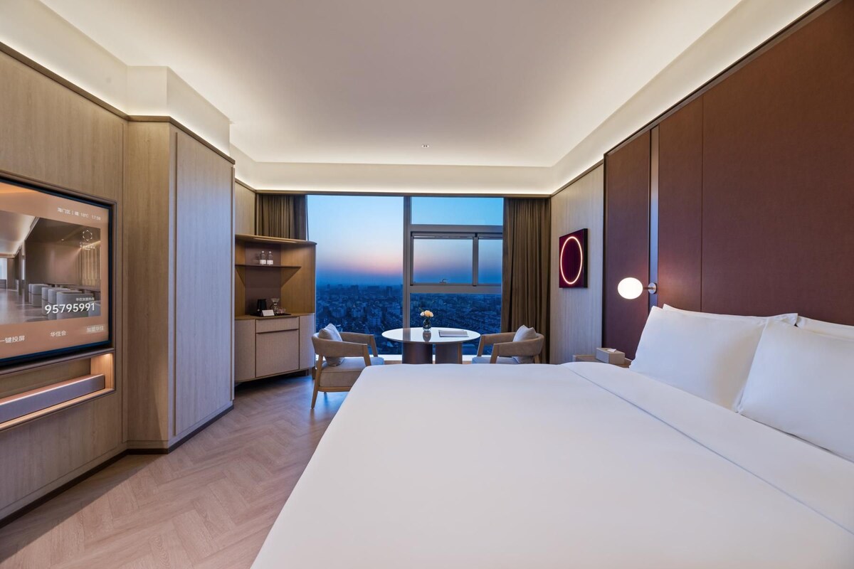 A hotel room with a large bed, a desk, a chair, a small table with a vase, and a view of the cityscape.