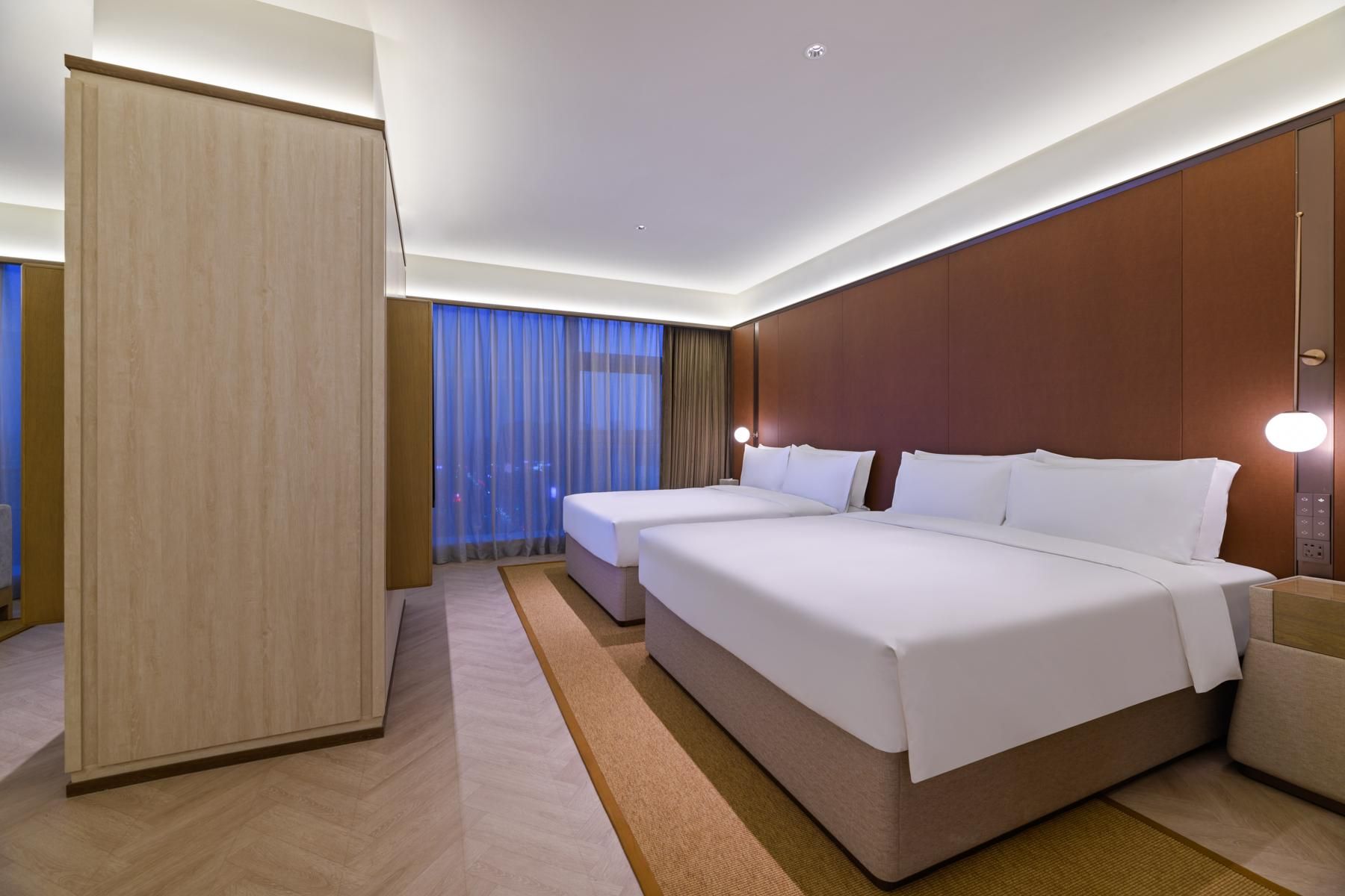 A modern hotel room with two beds, a large wardrobe, and a view of the city at night.