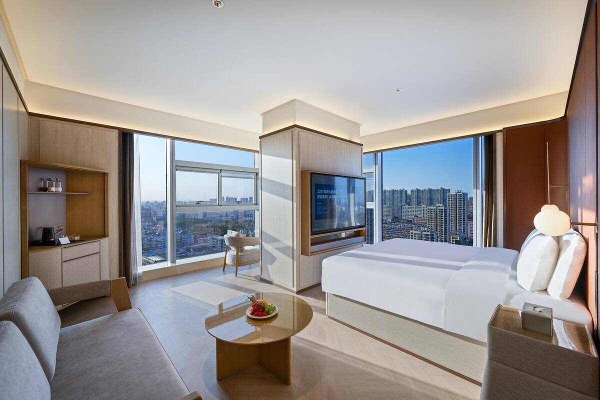 A modern hotel room with a large bed, a TV, a sofa, a coffee table, and a city view.