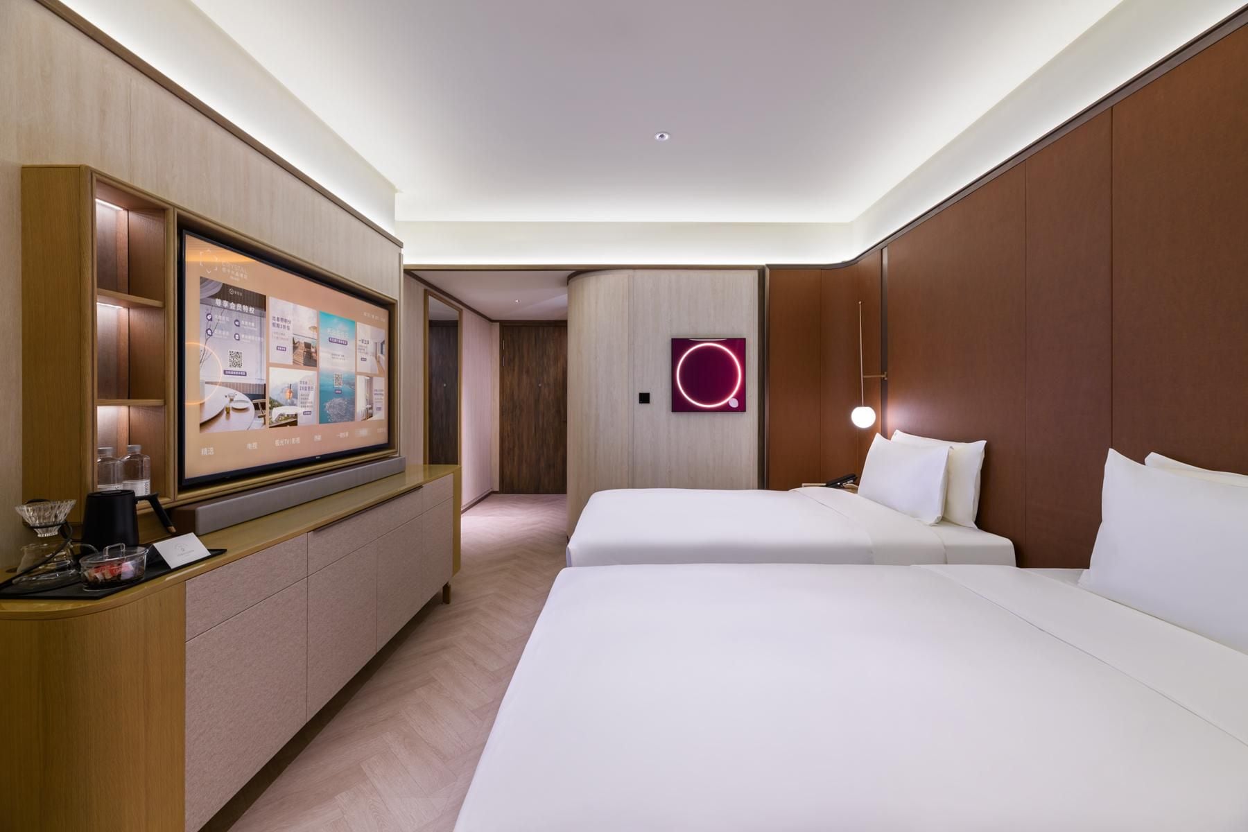 A hotel room with a large digital display, two beds, a desk with a coffee maker, and a wall-mounted light fixture.