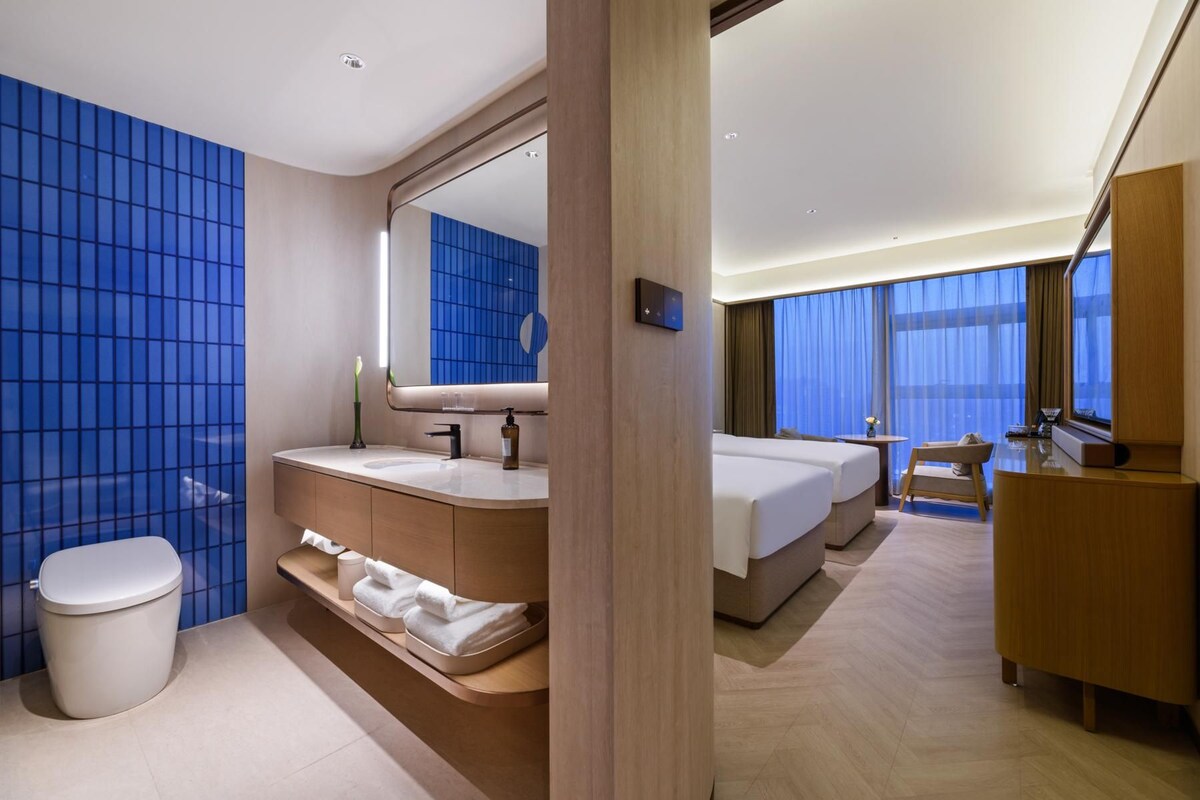 A modern hotel room with a large bed, a bathroom with a vanity, and a view of the city through large windows.