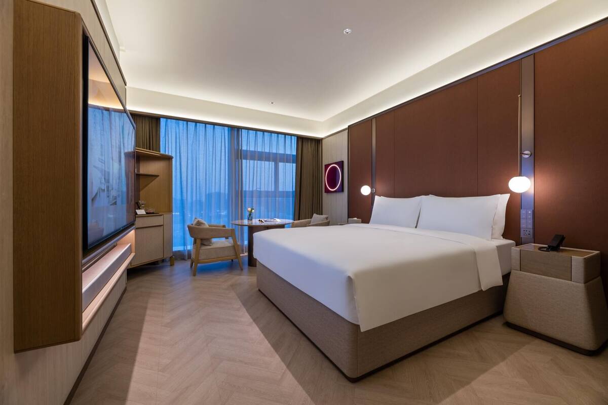 A modern hotel room with a large bed, a desk, and a chair. There is a TV on the left side and a wall-mounted light fixture above the bed.